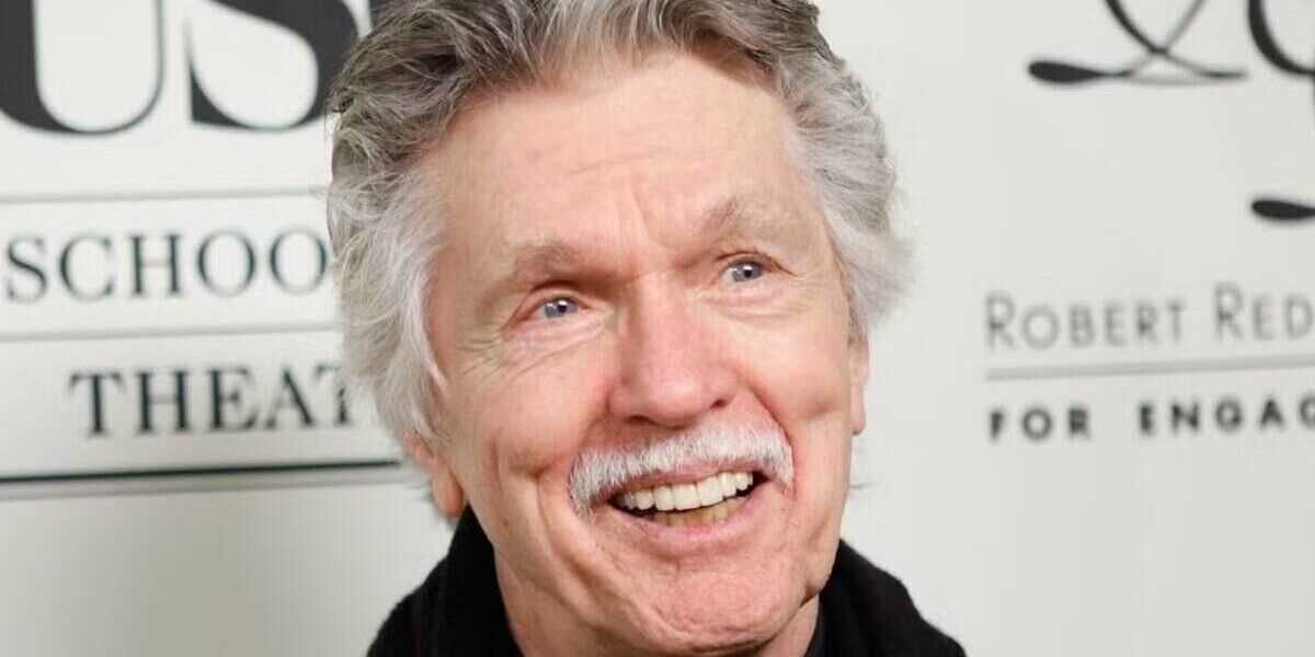Tom Skerritt, de cabello y bigote canosos, sonríe ante un panel con letras de un acto público