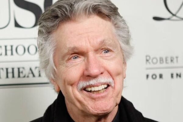 Tom Skerritt, de cabello y bigote canosos, sonríe ante un panel con letras de un acto público