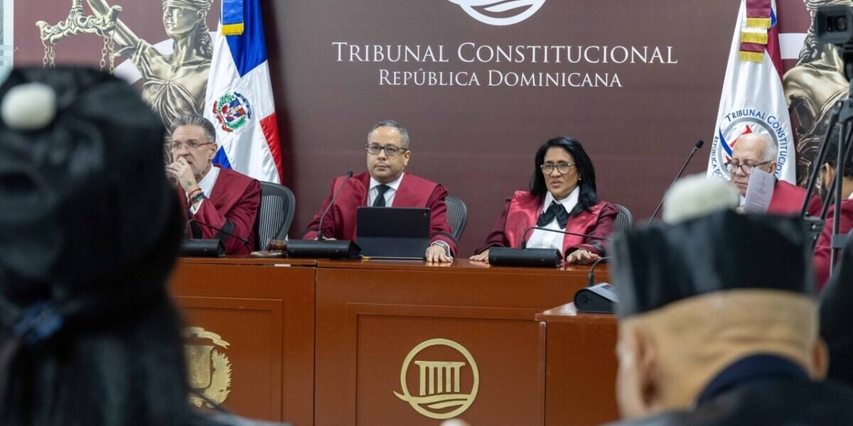 Jueces del Tribunal Constitucional con togas granate durante una audiencia, ante un fondo con el nombre de la institución y banderas dominicanas