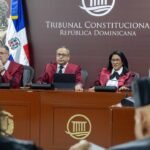 Jueces del Tribunal Constitucional con togas granate durante una audiencia, ante un fondo con el nombre de la institución y banderas dominicanas