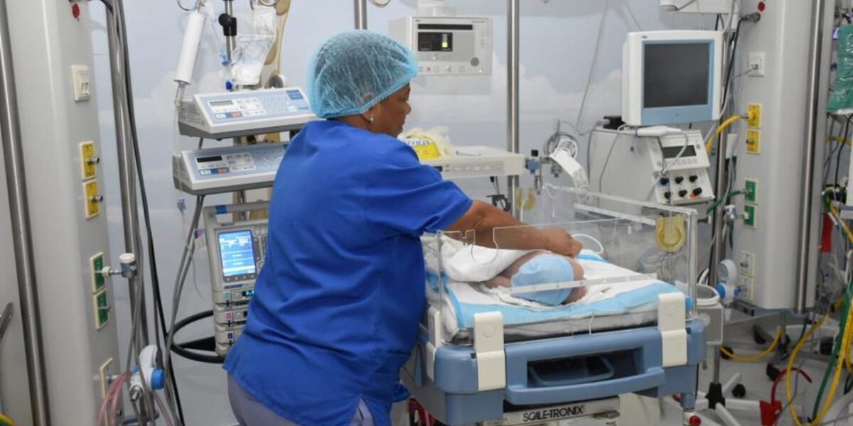 Una enfermera con uniforme azul y gorro atiende a un bebé en una cuna de cuidados intensivos rodeada de monitores y equipos médicos