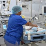 Una enfermera con uniforme azul y gorro atiende a un bebé en una cuna de cuidados intensivos rodeada de monitores y equipos médicos