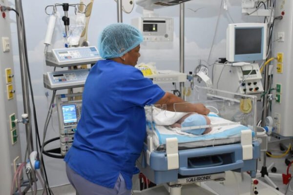 Una enfermera con uniforme azul y gorro atiende a un bebé en una cuna de cuidados intensivos rodeada de monitores y equipos médicos