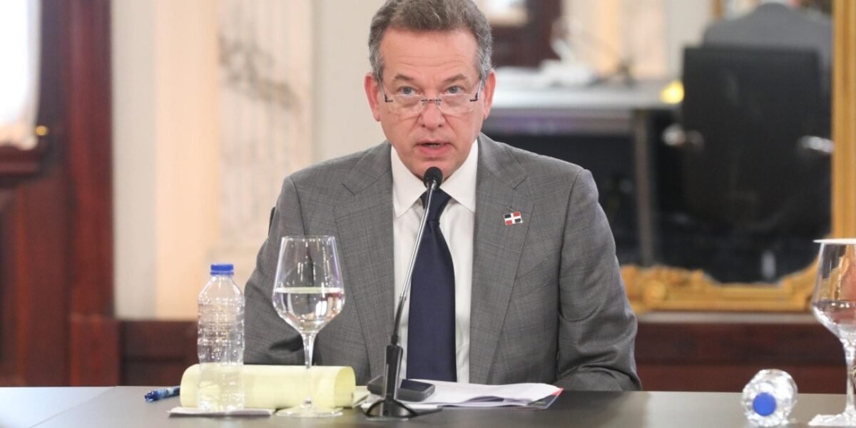 Víctor Bisonó, con traje gris, corbata azul y un pin de la bandera dominicana, habla ante un micrófono sentado a una mesa