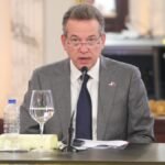 Víctor Bisonó, con traje gris, corbata azul y un pin de la bandera dominicana, habla ante un micrófono sentado a una mesa