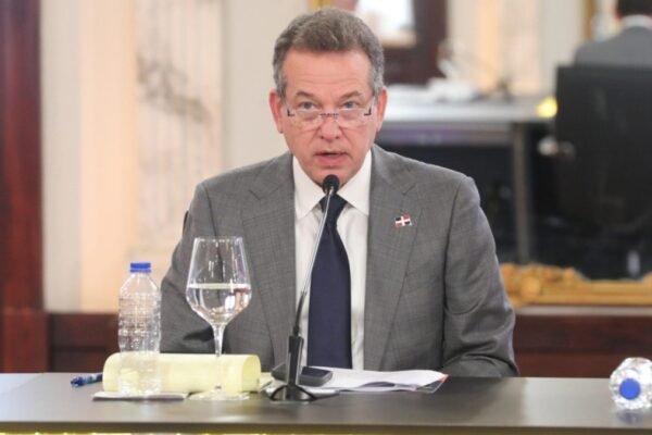 Víctor Bisonó, con traje gris, corbata azul y un pin de la bandera dominicana, habla ante un micrófono sentado a una mesa