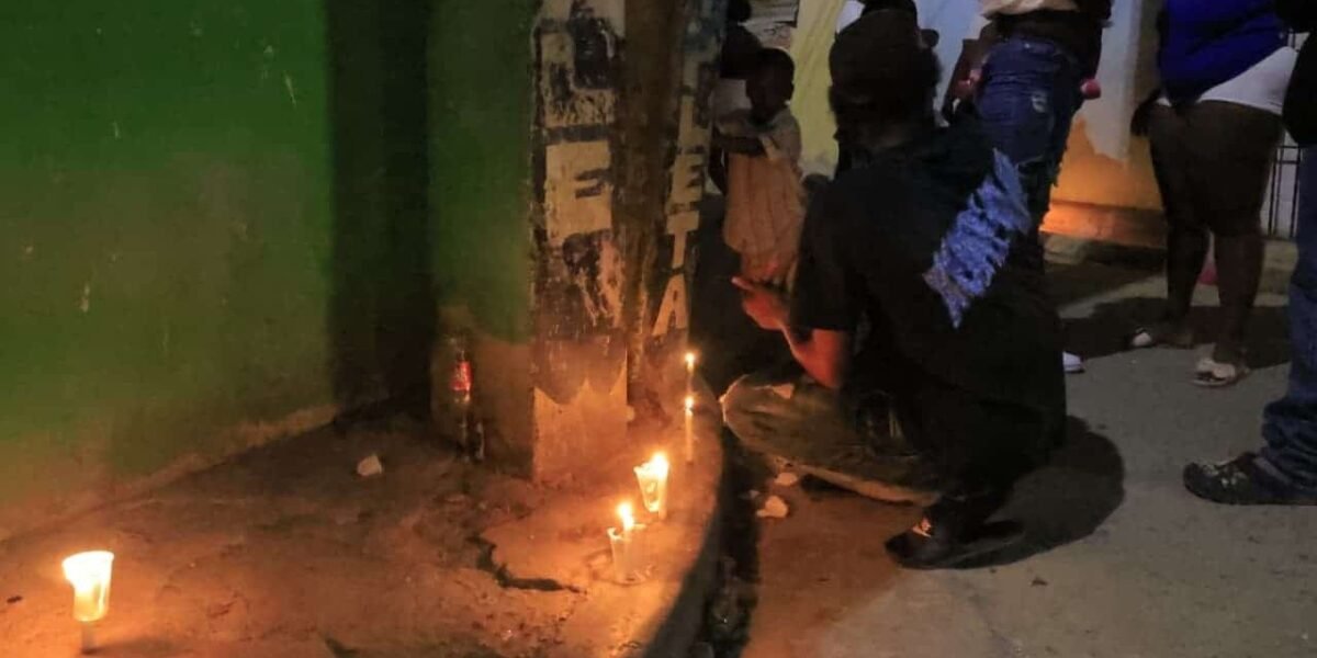 Velas encendidas en la acera junto a un poste, con un hombre agachado y vecinos de pie alrededor durante una vigilia nocturna