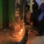 Velas encendidas en la acera junto a un poste, con un hombre agachado y vecinos de pie alrededor durante una vigilia nocturna
