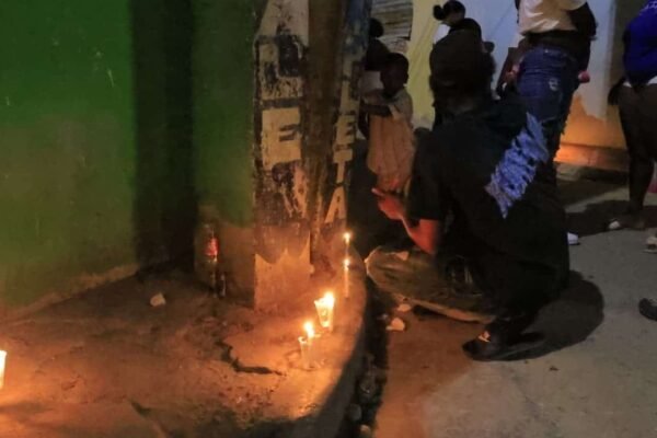 Velas encendidas en la acera junto a un poste, con un hombre agachado y vecinos de pie alrededor durante una vigilia nocturna