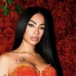 Retrato de la artista Yailin La Más Viral, con el cabello largo y negro y un corpiño naranja, ante un fondo de rosas rojas