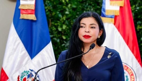 La procuradora general Yeni Berenice Reynoso, con blusa azul marino, habla ante un micrófono frente a dos banderas dominicanas.