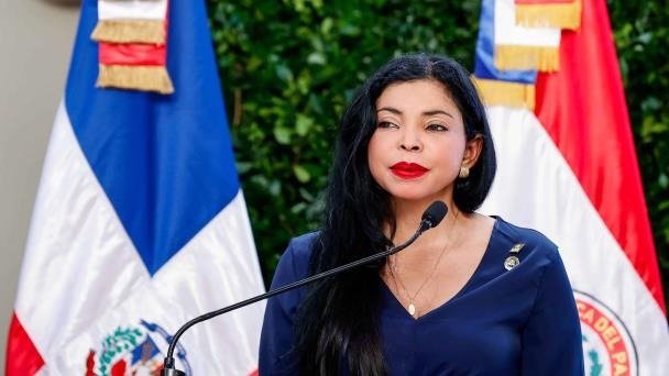 La procuradora general Yeni Berenice Reynoso, con blusa azul marino, habla ante un micrófono frente a dos banderas dominicanas.