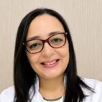 Ylsa Quero Cabrera, con gafas y bata blanca, sonríe ante una pared beige