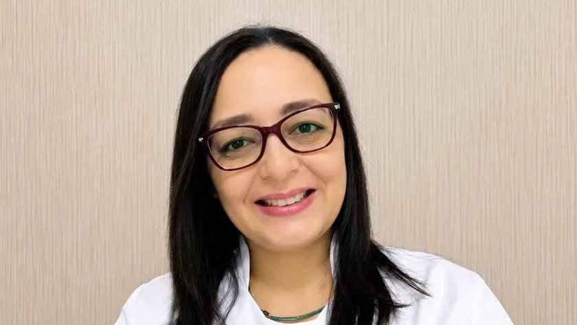 Ylsa Quero Cabrera, con gafas y bata blanca, sonríe ante una pared beige